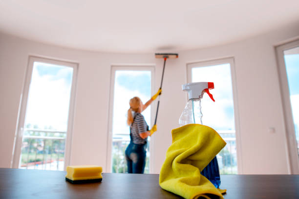 Yellow cleaning rag and sponge with spraying bottle on desk with blurred housewife cleaning the ceiling in the background. House cleaning concept. Copy space